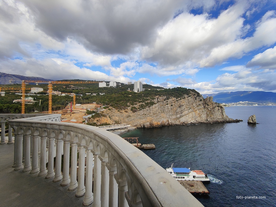 Вид со смотровой площадки с балюстрадой у западной стены замка "Ласточкино гнездо" на вершине Аврориной скалы на побережье мыса Ай-Тодор с действующей и строящейся инфраструктурой и его восточный (Лимен-Бурун) отрог с отдельной маленькой скалой Парус